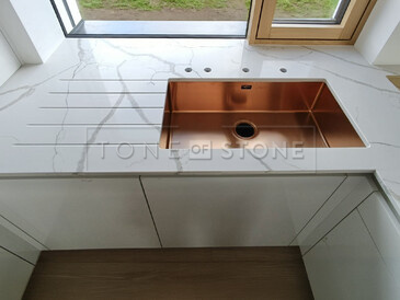 Realizace pracovní desky z technistone Crystal Calacatta Silva zahrnující měděný dřez se spodní montáží, vyfrézované odkapové drážky a výřezy pro baterii a dávkovač saponátu. Kamenný parapet je součástí kuchyňské desky. Zakázková výroba TONE OF STONE. Realizace pracovní desky z technistone Crystal Calacatta Silva zahrnující měděný dřez se spodní montáží, vyfrézované odkapové drážky a výřezy pro baterii a dávkovač saponátu. Kamenný parapet je součástí kuchyňské desky. Zakázková výroba TONE OF STONE.
